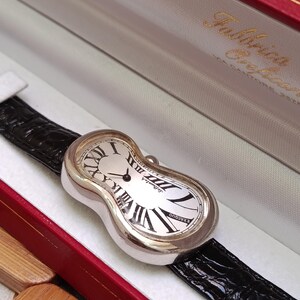 May include: A silver-toned wristwatch with a unique, melting design. The watch face is white with black Roman numerals and hands. It has a black leather band and is presented in a red and white box.