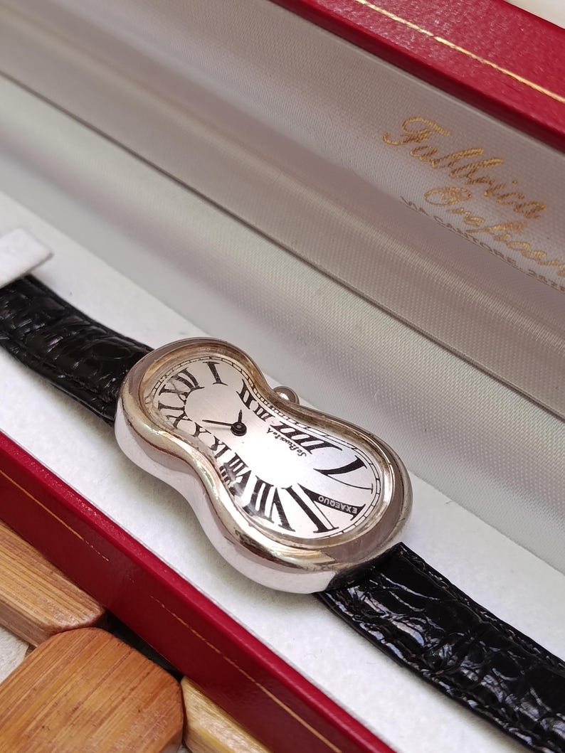 May include: A silver-toned wristwatch with a unique, melting design. The watch face is white with black Roman numerals and hands. It has a black leather band and is presented in a red and white box.