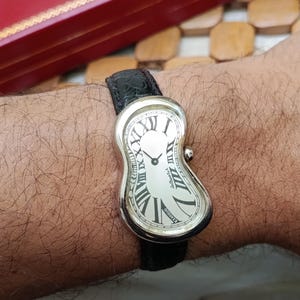 May include: A silver-toned wristwatch with a melting, curved design. The watch face is white with black Roman numerals and hands. The watch has a black leather band.
