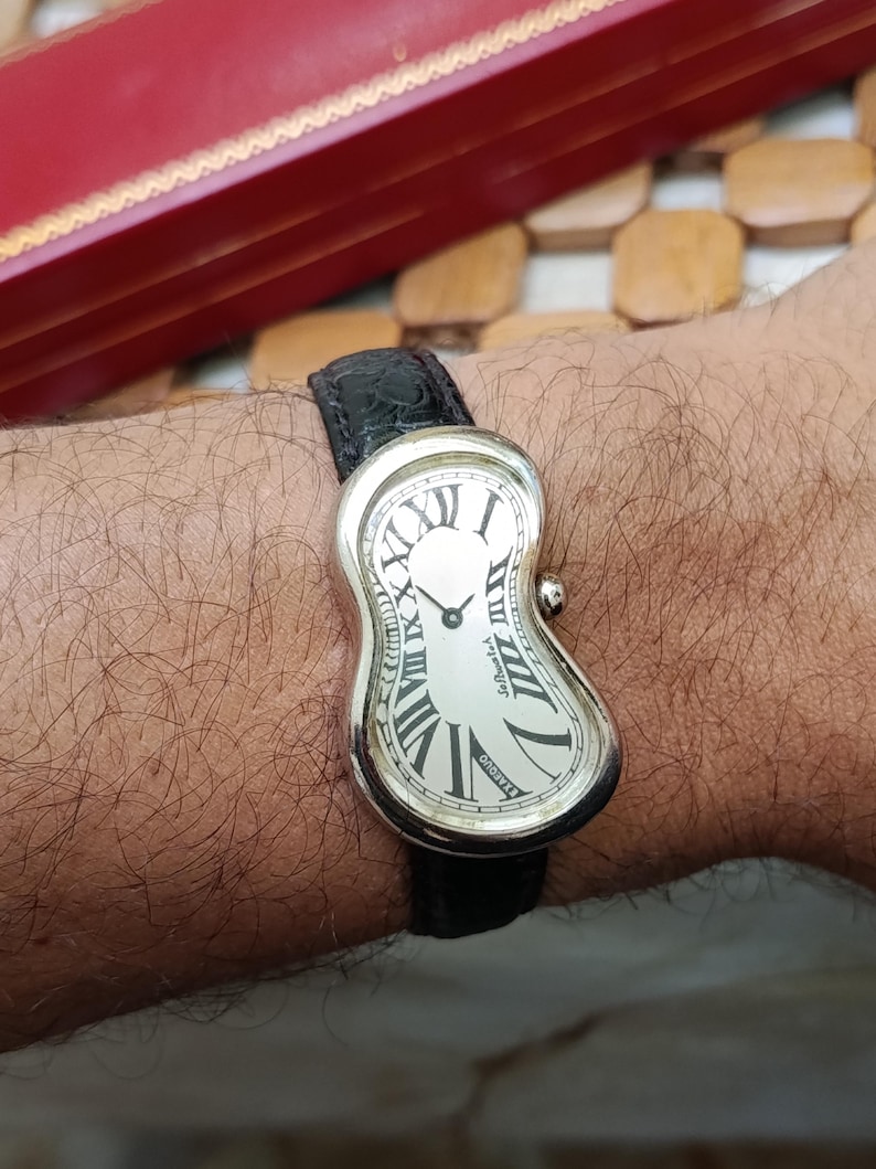 May include: A silver-toned wristwatch with a melting, curved design. The watch face is white with black Roman numerals and hands. The watch has a black leather band.