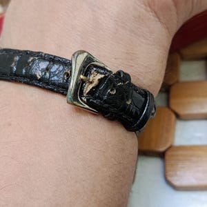 May include: Close-up of a black leather watch strap with a silver buckle. The strap shows signs of wear and tear. The watch strap is attached to a wrist.
