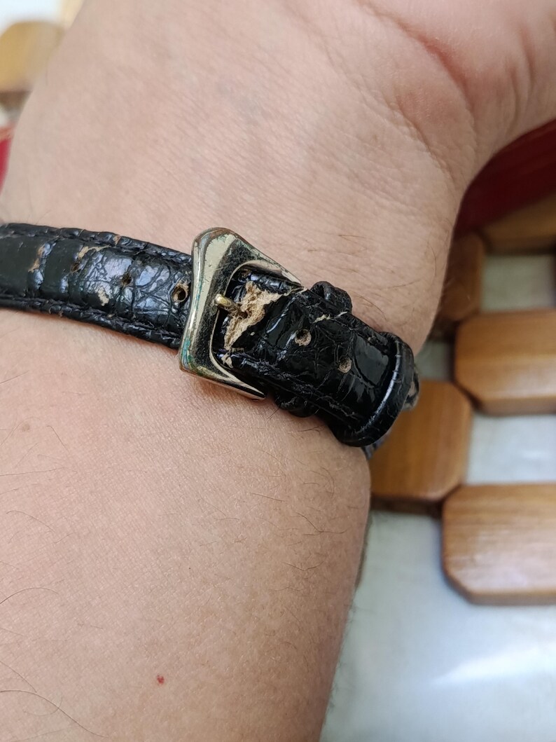 May include: Close-up of a black leather watch strap with a silver buckle. The strap shows signs of wear and tear. The watch strap is attached to a wrist.
