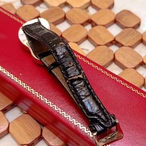May include: A silver wristwatch with a black leather band is displayed on a red box. The watch band is textured and has a gold-colored buckle. The watch is set against a background of wooden tiles.
