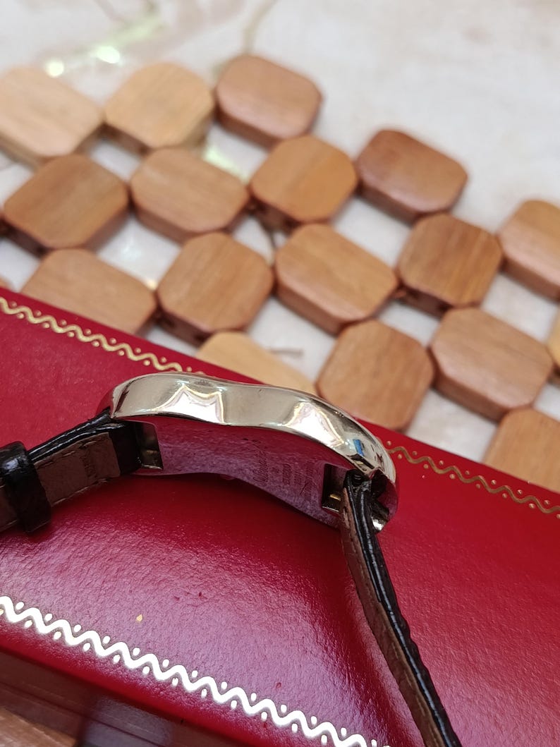 May include: A silver wristwatch with a black leather band. The watch is set on a red surface with a decorative border. The background features a pattern of small, wooden, square-shaped objects.