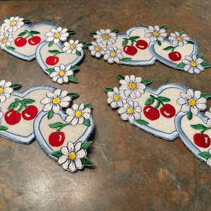 May include: Six embroidered iron-on patches featuring a pair of red cherries with green stems and leaves, surrounded by white daisies with yellow centers, on a blue heart-shaped background.