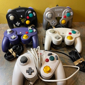 May include: Five Nintendo GameCube controllers in different colors: black, silver, purple, white, and white with red, green, and yellow buttons. The controllers are all wired and have the Nintendo GameCube logo on them.