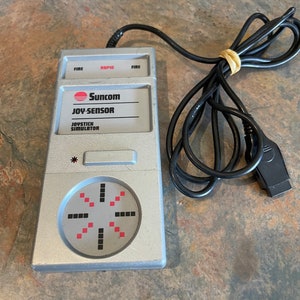 May include: A silver Suncom Joy-Sensor joystick simulator with a red and black directional pad and buttons labeled "Fire", "Rapid", and "Fire".