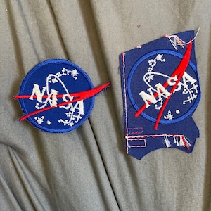 May include: Two NASA patches, one blue and white with a red rocket, the other blue and white with a red rocket and a frayed edge.