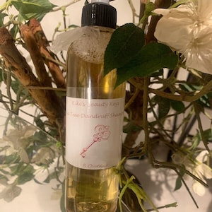 May include: A clear glass bottle with a white label that reads "Kiki's Beauty Keys Tea Tree Dandruff Shampoo" and "8 Ounces". The bottle is sitting on a table with a green plant in the background.