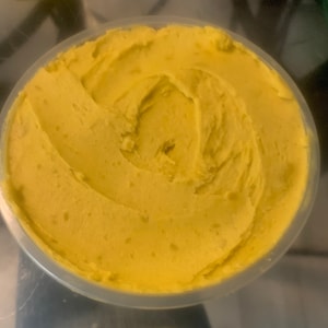 May include: A container of yellow-colored cream with a smooth, swirled texture. The cream is in a clear, round plastic container. The product appears to be a skincare or cosmetic item.