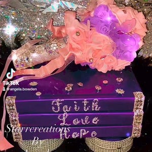 May include: A stack of three purple books decorated with rhinestone trim and the words Faith, Love, and Hope in crystal lettering. A bouquet of peach and purple fabric roses with butterfly accents and a rhinestone-encrusted handle rests on top.