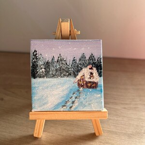 Peut inclure: Une petite peinture sur toile représentant une scène d'hiver enneigée avec une petite cabane au loin. La peinture est sur un chevalet en bois.