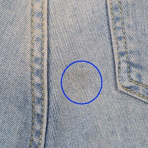 May include: Close-up of light blue denim fabric with a faint yellow stain. The fabric has a subtle woven texture.