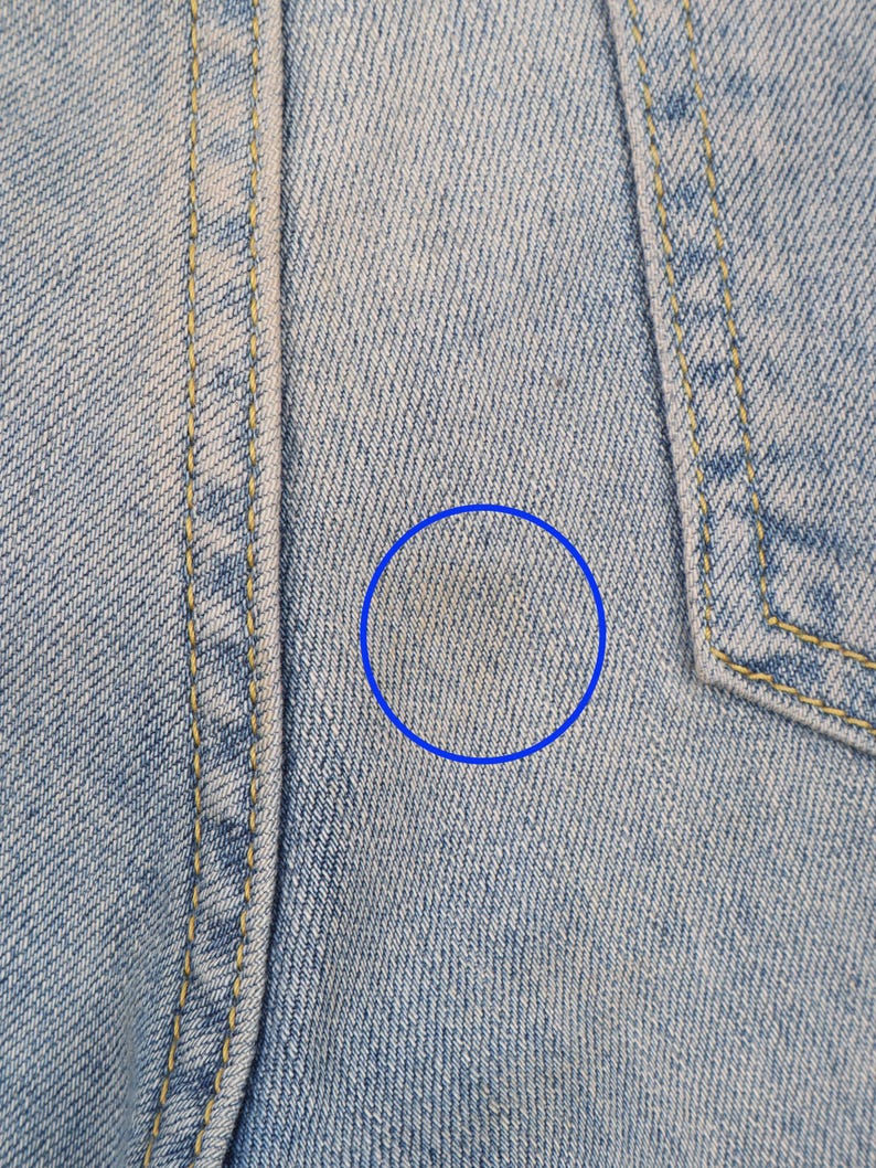 May include: Close-up of light blue denim fabric with a faint yellow stain. The fabric has a subtle woven texture.