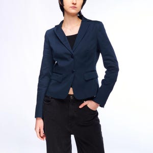 Puede incluir: Una chaqueta azul oscuro con cierre de un solo botón y dos bolsillos delanteros. La chaqueta se lleva sobre una camiseta negra y unos vaqueros oscuros.