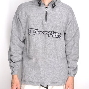 May include: A gray Champion sweatshirt with a black zipper and a large embroidered Champion logo on the front. The logo is in a white and silver color scheme and says "Champion U.S.A."