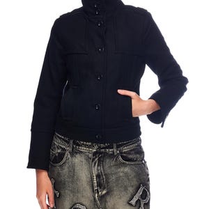 May include: A black jacket with a high collar and button closure. The jacket has two front pockets and long sleeves. The person is wearing acid wash jeans with letter and cross designs.