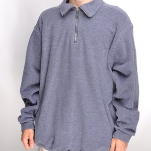 May include: A blue-grey pullover sweatshirt with a collared neckline and a silver zipper. The long-sleeved top has a drawstring at the waist. The model is wearing light-colored shorts.