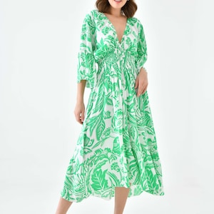 Puede incluir: Un vestido midi blanco con estampado floral verde. El vestido tiene un escote en V, mangas 3/4 y una cintura fruncida. La modelo lleva sandalias blancas.