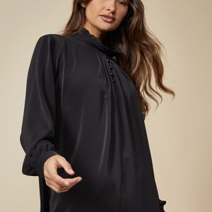 Puede incluir: Una blusa negra de manga larga con cuello alto y una fila de peque&ntilde;os botones negros en la parte delantera.