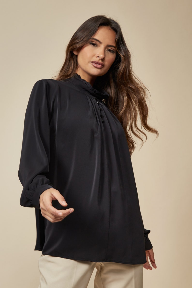Puede incluir: Una blusa negra de manga larga con cuello alto y una fila de peque&ntilde;os botones negros en la parte delantera.