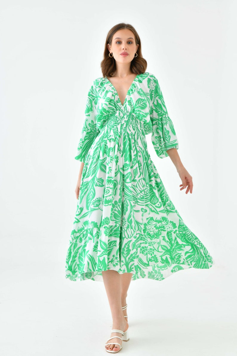 Puede incluir: Un vestido midi blanco con estampado floral verde. El vestido tiene un escote en V, mangas 3/4 y una cintura ajustada. La modelo lleva sandalias blancas.