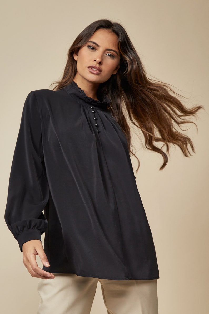 Puede incluir: Una blusa negra de manga larga con cuello alto y una fila de peque&ntilde;os botones negros en la parte delantera.