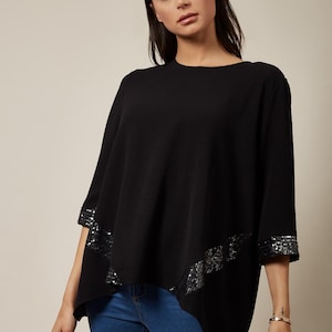 Puede incluir: Top negro con detalles de lentejuelas en las mangas y el bajo. El top tiene un diseño holgado y se combina con vaqueros azules. Las mangas son de tres cuartos.