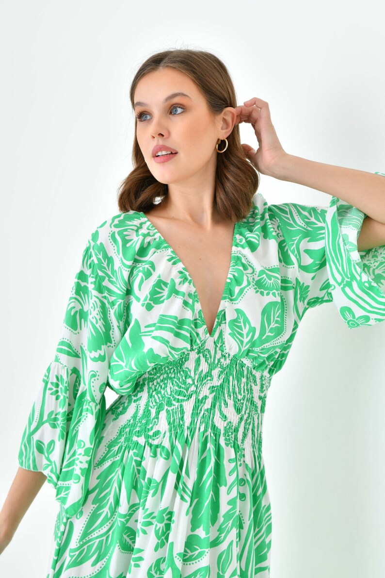 Puede incluir: Un vestido blanco con estampado floral verde y un escote en V profundo. El vestido tiene una cintura fruncida y mangas cortas abullonadas.
