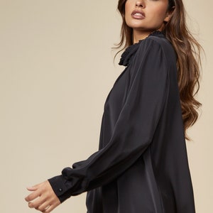 Puede incluir: Una blusa negra de manga larga con cuello fruncido. La blusa tiene un corte holgado y est&aacute; hecha de una tela suave y sedosa.