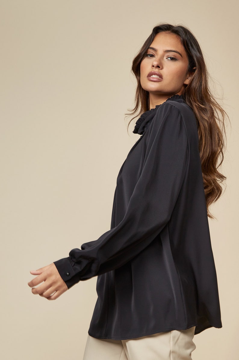 Puede incluir: Una blusa negra de manga larga con cuello fruncido. La blusa tiene un corte holgado y est&aacute; hecha de una tela suave y sedosa.