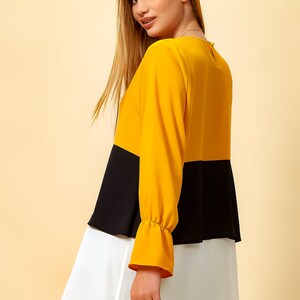 May include: A three-toned blouse with a yellow top, black middle, and white bottom. The blouse has long sleeves with a ruffled cuff and a round neckline.