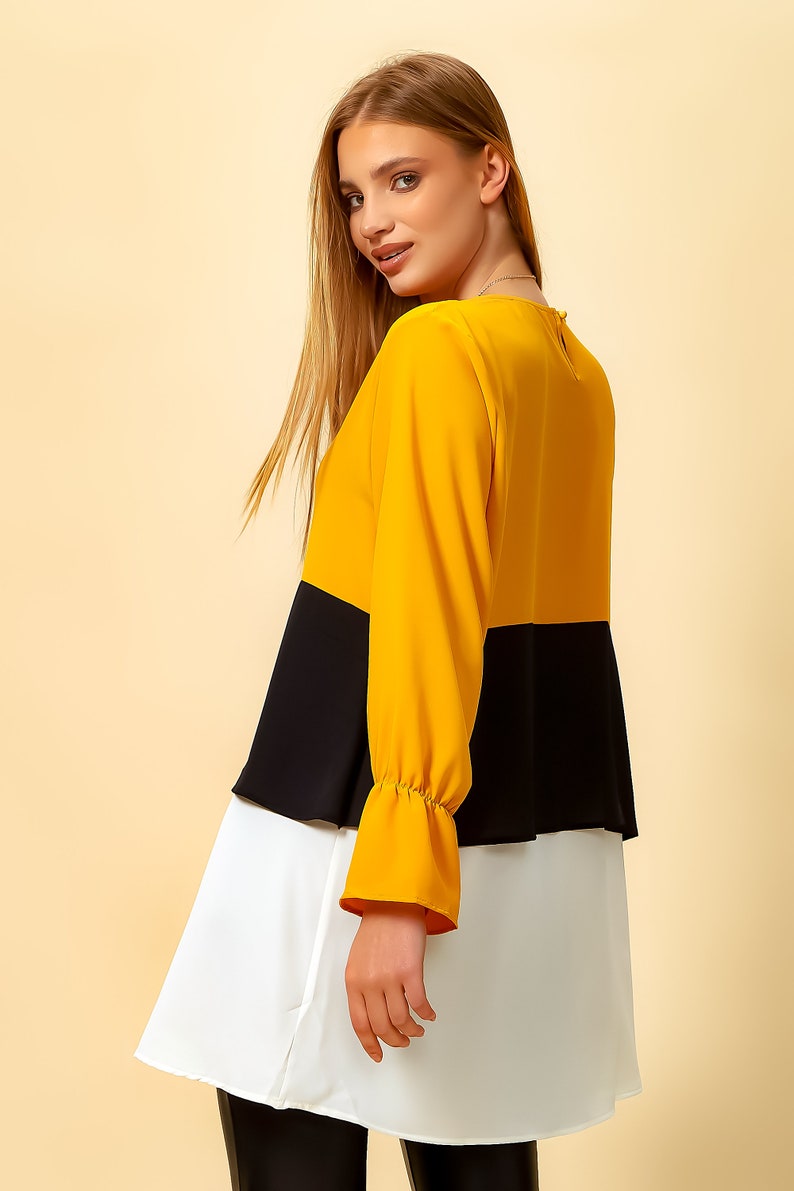 May include: A three-toned blouse with a yellow top, black middle, and white bottom. The blouse has long sleeves with a ruffled cuff and a round neckline.