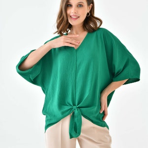 May include: A green, loose-fitting,  top with a V-neck and a knot at the waist. The top is made of a soft, textured fabric. The model is wearing the top with beige pants.