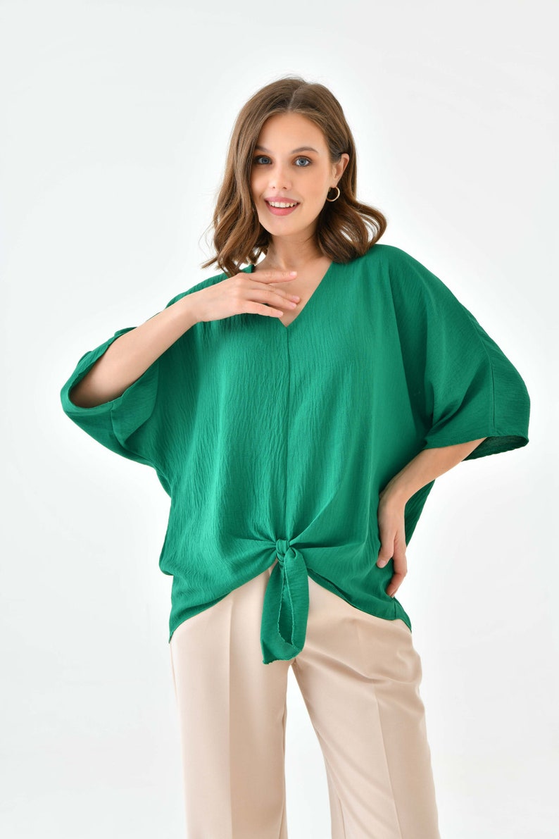 May include: A green, loose-fitting,  top with a V-neck and a knot at the waist. The top is made of a soft, textured fabric. The model is wearing the top with beige pants.