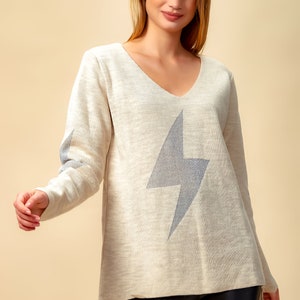 May include: A beige sweater with a silver glitter lightning bolt design. The sweater has a v-neck and long sleeves.