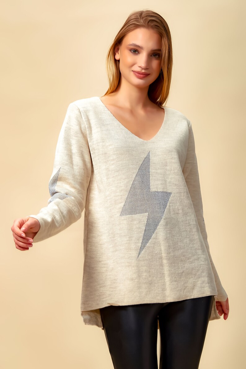 May include: A beige sweater with a silver glitter lightning bolt design. The sweater has a v-neck and long sleeves.
