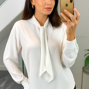 Op de afbeelding: Witte blouse met lange mouwen en een strik aan de hals. De vrouw draagt gouden oorringen en houdt een gouden telefoon vast. Ze draagt een zwarte broek.