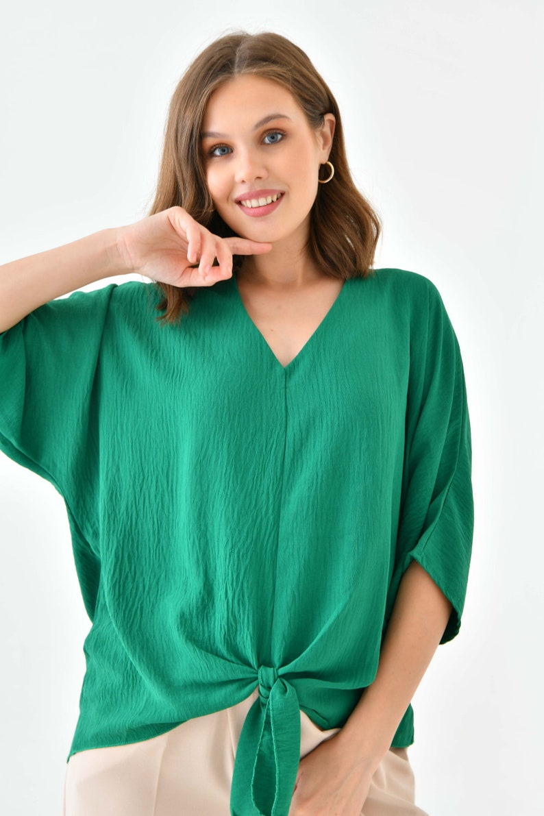 May include: A green, short-sleeved, v-neck blouse with a knotted front. The blouse is made of a textured fabric.