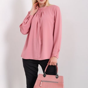 K&ouml;nnte beinhalten: Eine rosa Bluse mit langen &Auml;rmeln, einem R&uuml;schenkragen und Kn&ouml;pfen. Die Bluse hat eine gefaltete Vorderseite und wird mit einer schwarzen Hose kombiniert. Die Person tr&auml;gt eine rosa Handtasche aus Krokodilleder.