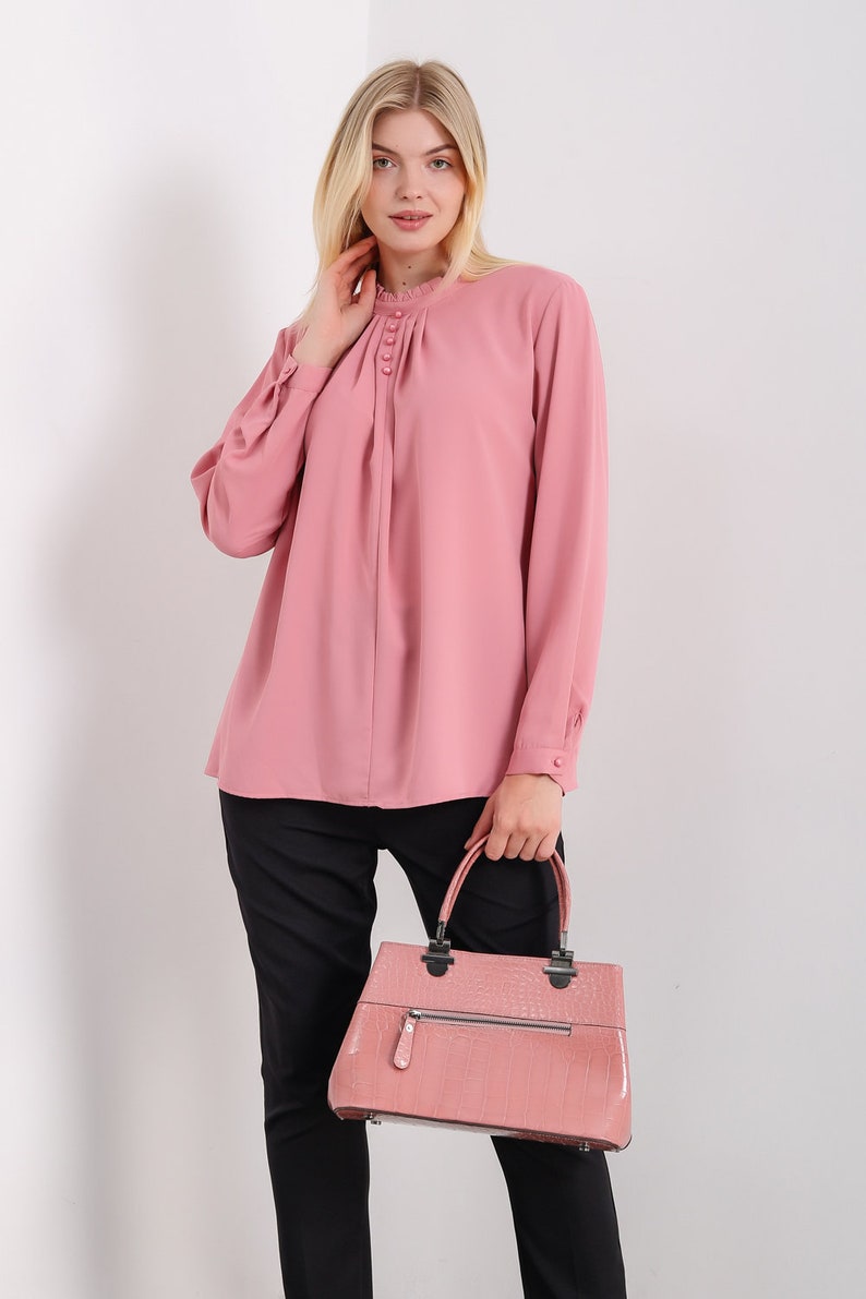 K&ouml;nnte beinhalten: Eine rosa Bluse mit langen &Auml;rmeln, einem R&uuml;schenkragen und Kn&ouml;pfen. Die Bluse hat eine gefaltete Vorderseite und wird mit einer schwarzen Hose kombiniert. Die Person tr&auml;gt eine rosa Handtasche aus Krokodilleder.