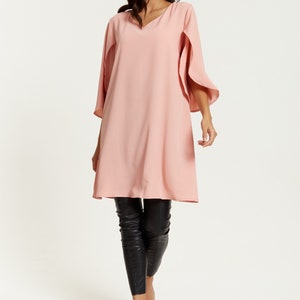 Op de afbeelding: Een roze, lange mouwen tuniek met een v-hals en een cape-achtige mouw detail. De tuniek wordt gedragen met een zwarte legging en zwarte sandalen met hak.