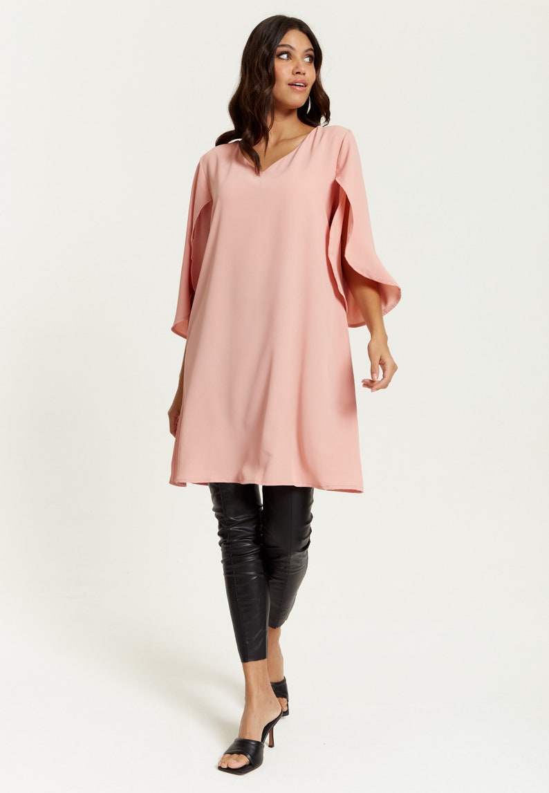 Op de afbeelding: Een roze, lange mouwen tuniek met een v-hals en een cape-achtige mouw detail. De tuniek wordt gedragen met een zwarte legging en zwarte sandalen met hak.