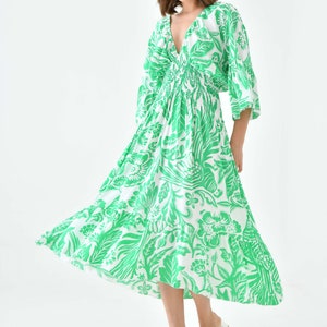 Puede incluir: Un vestido midi blanco con estampado floral verde. El vestido tiene un escote en V, mangas 3/4 y una cintura fruncida. La modelo lleva sandalias blancas.