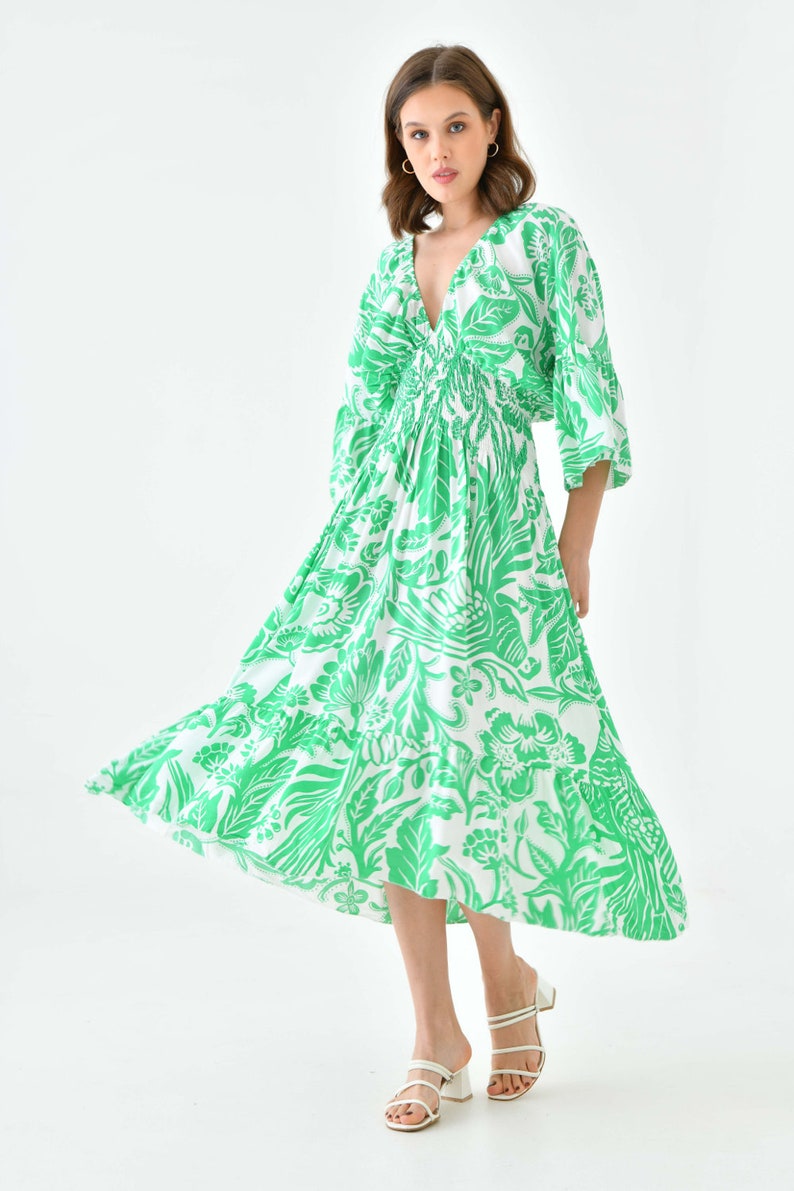Puede incluir: Un vestido midi blanco con estampado floral verde. El vestido tiene un escote en V, mangas 3/4 y una cintura fruncida. La modelo lleva sandalias blancas.