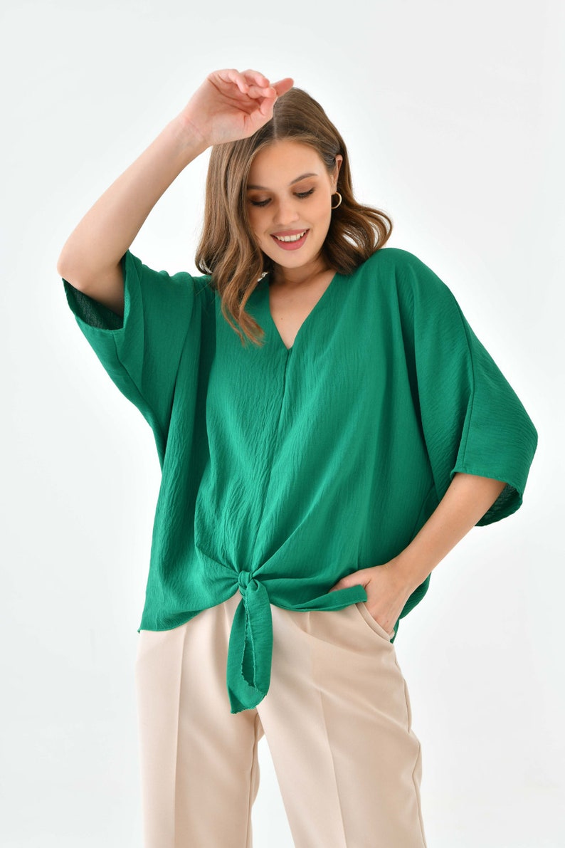 May include: A green, loose-fitting, short-sleeved blouse with a knotted front. The blouse is made of a textured fabric and has a v-neck.