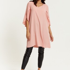 Op de afbeelding: Een roze tuniek met lange mouwen, een V-hals en fladderende mouwen. De tuniek wordt gedragen met een zwarte legging en zwarte sandalen met hak.