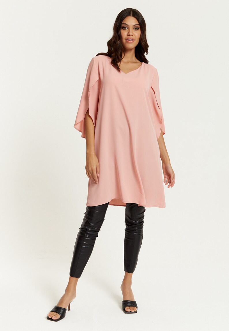 Op de afbeelding: Een roze tuniek met lange mouwen, een V-hals en fladderende mouwen. De tuniek wordt gedragen met een zwarte legging en zwarte sandalen met hak.