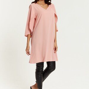 Op de afbeelding: Een roze lange mouwen tuniek met een v-hals en fladderende mouwen. De tuniek is gecombineerd met een zwarte legging en zwarte sandalen met hak.