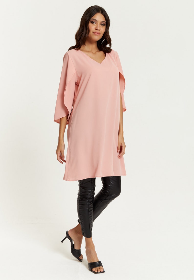 Op de afbeelding: Een roze lange mouwen tuniek met een v-hals en fladderende mouwen. De tuniek is gecombineerd met een zwarte legging en zwarte sandalen met hak.